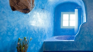 A room with a blue-tiled floor, a large rock, a window, and a tiled wall. - Vallas Apartments & Villas (Santorini)