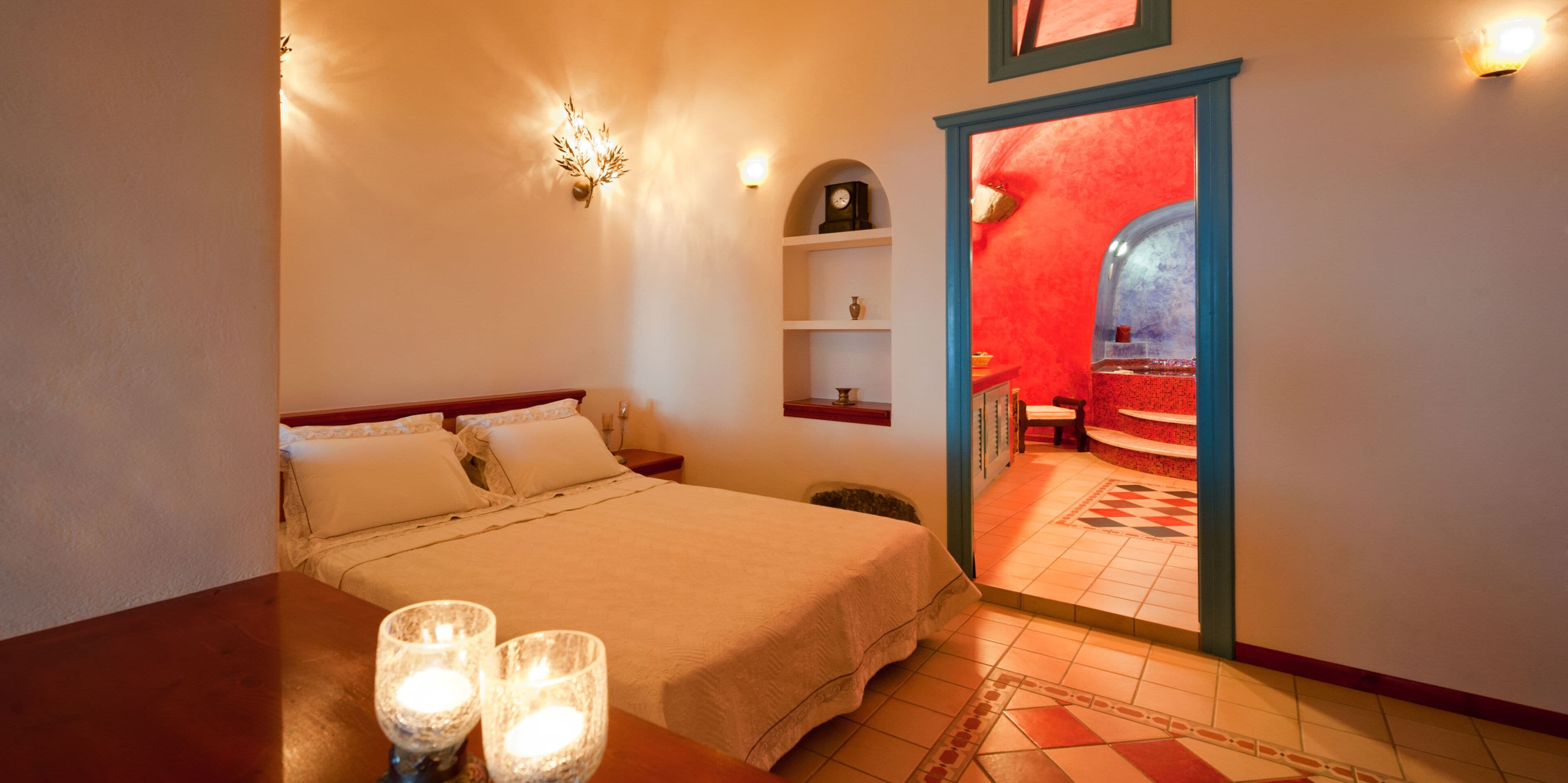 A bedroom with a bed, bedside table, and a nightstand. There is a small shelf with a clock and a vase. The room has a red accent wall and a tiled floor.