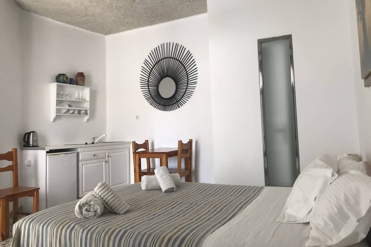 Une chambre à coucher comprenant un lit, une chaise en bois, un bureau avec une bouilloire, un miroir et une étagère murale où sont rangés de la vaisselle.