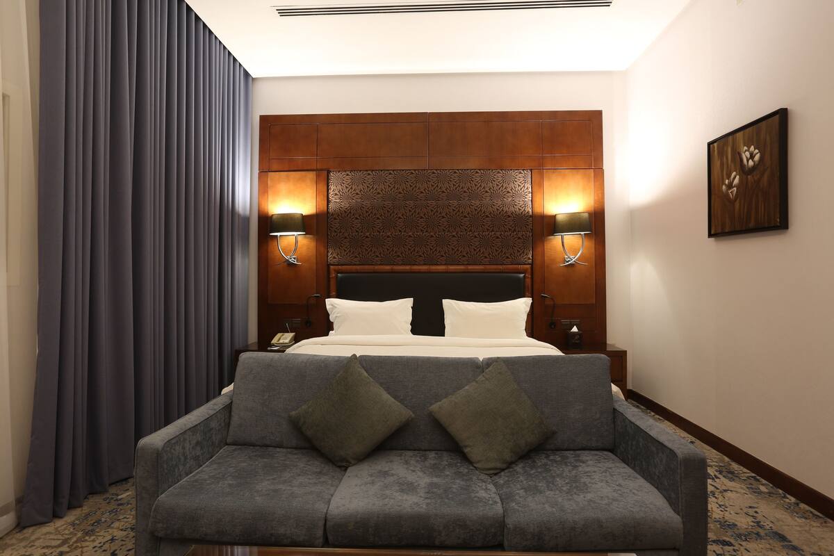 Quarto de hotel com cama, duas luminárias de cabeceira, sofá com almofadas, mesa de cabeceira, obra de arte na parede e tapete estampado.