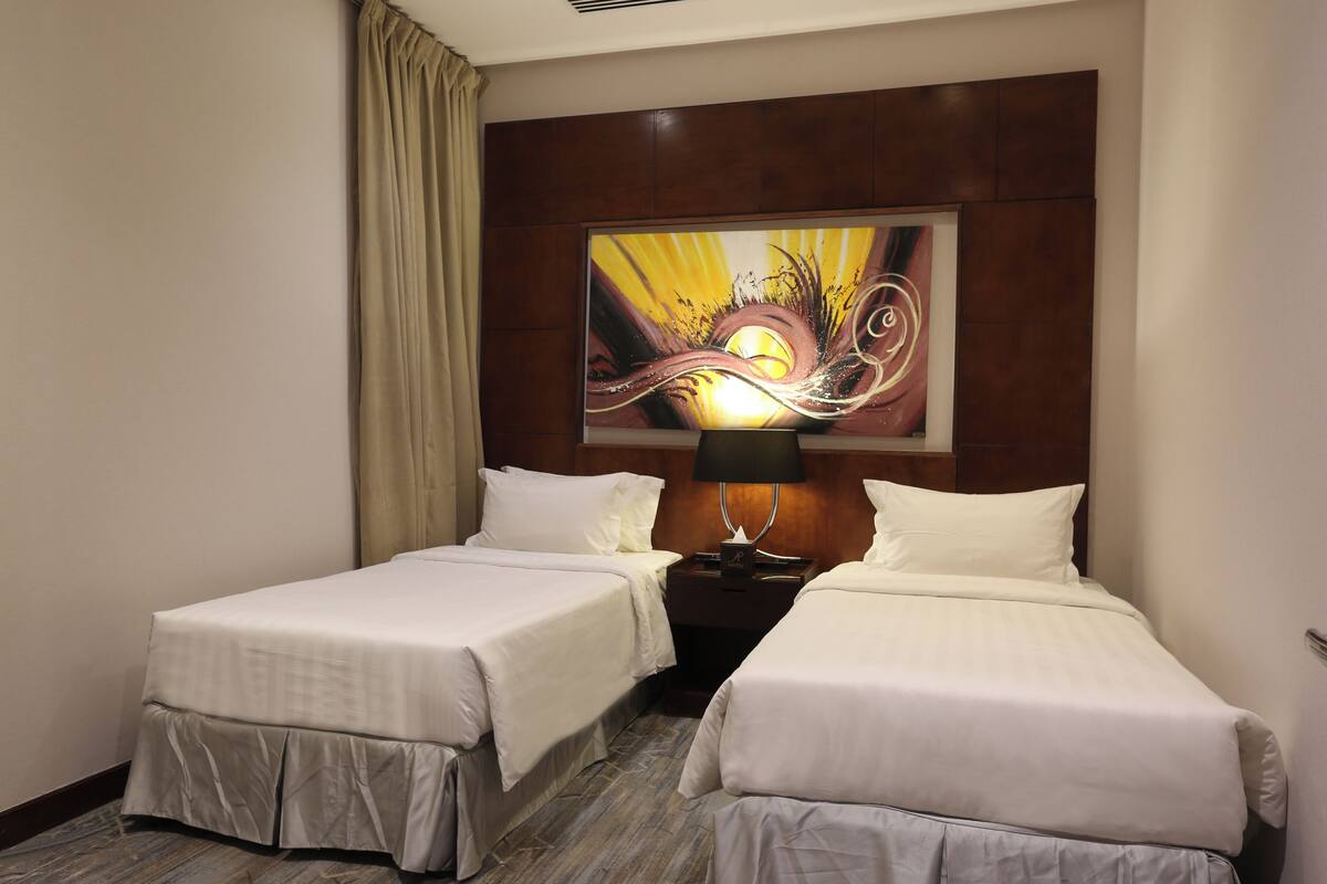 Quarto de hotel com duas camas, uma mesa de cabeceira, uma luminária e um quadro abstrato grande na parede.