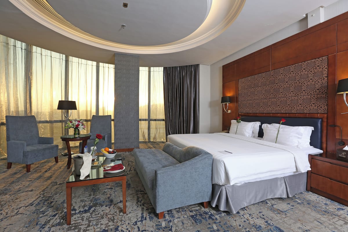 Une chambre d’hôtel avec un grand lit, deux fauteuils, une petite table ornée d’un vase de fleurs et une vue sur l’extérieur grâce à de grandes fenêtres.