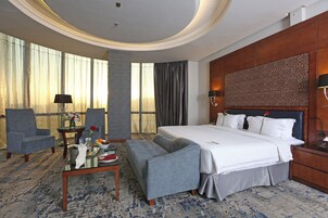 A hotel room with a large bed, two armchairs, a small table with a vase of flowers, and a view of the outdoors through large windows.