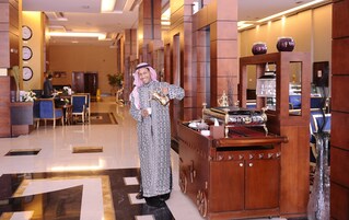 A man in traditional attire holding a teapot in a hotel lobby with a reception desk, chairs, and clocks on the wall.