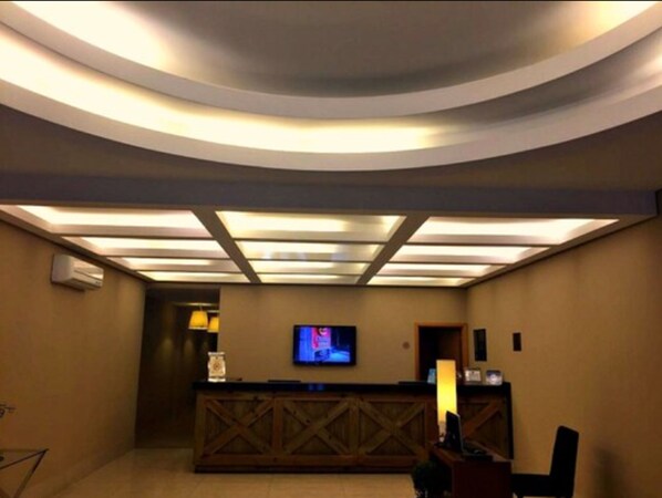 Zona de recepción de un hotel con un mostrador de madera, un televisor de pantalla plana montado en la pared e iluminación empotrada en el techo.