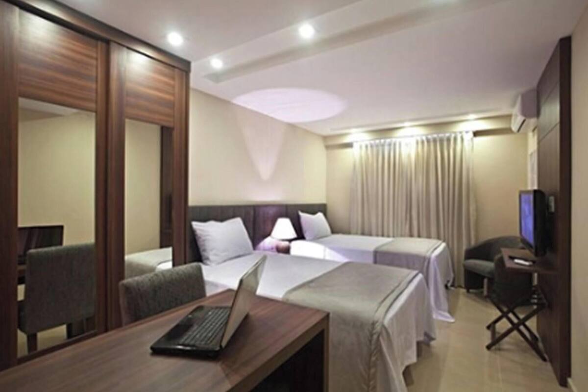 Ein Hotelzimmer mit einem Bett, einem Schreibtisch mit einem Laptop, einem Sessel, einem Fernseher und einem Vorhang.