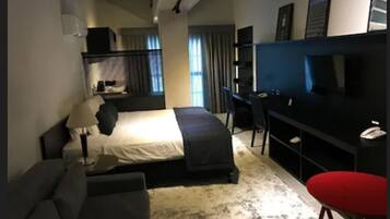 A hotel room with a bed, a desk, a chair, a TV, and a red stool.