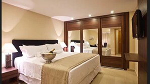A hotel room with a large bed, a bedside table with a lamp, a minibar, a TV mounted on the wall, and a sliding door closet. - Hotel Granja Brasil Resort (Petrópolis)