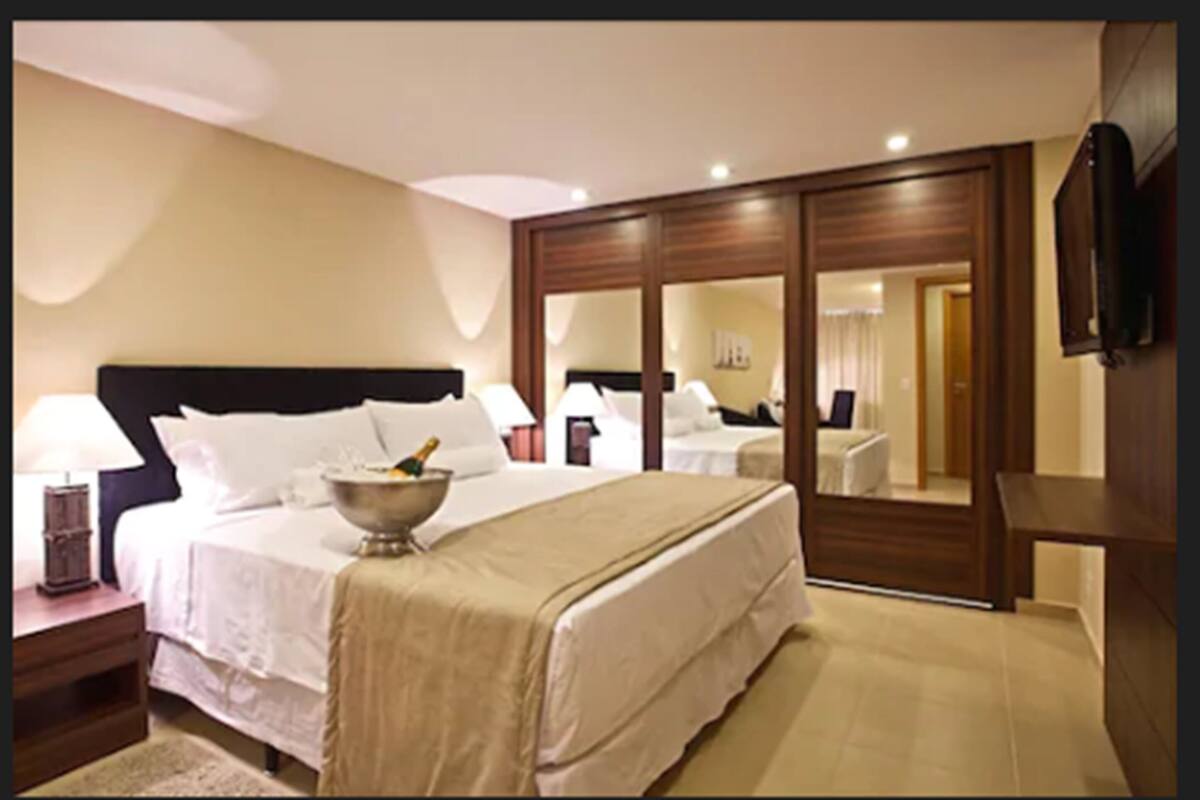 Ein Hotelzimmer mit einem großen Bett, einem Nachttisch mit Lampe, einer Minibar, einem an der Wand montierten Fernseher und einem Schiebetürenschrank.
