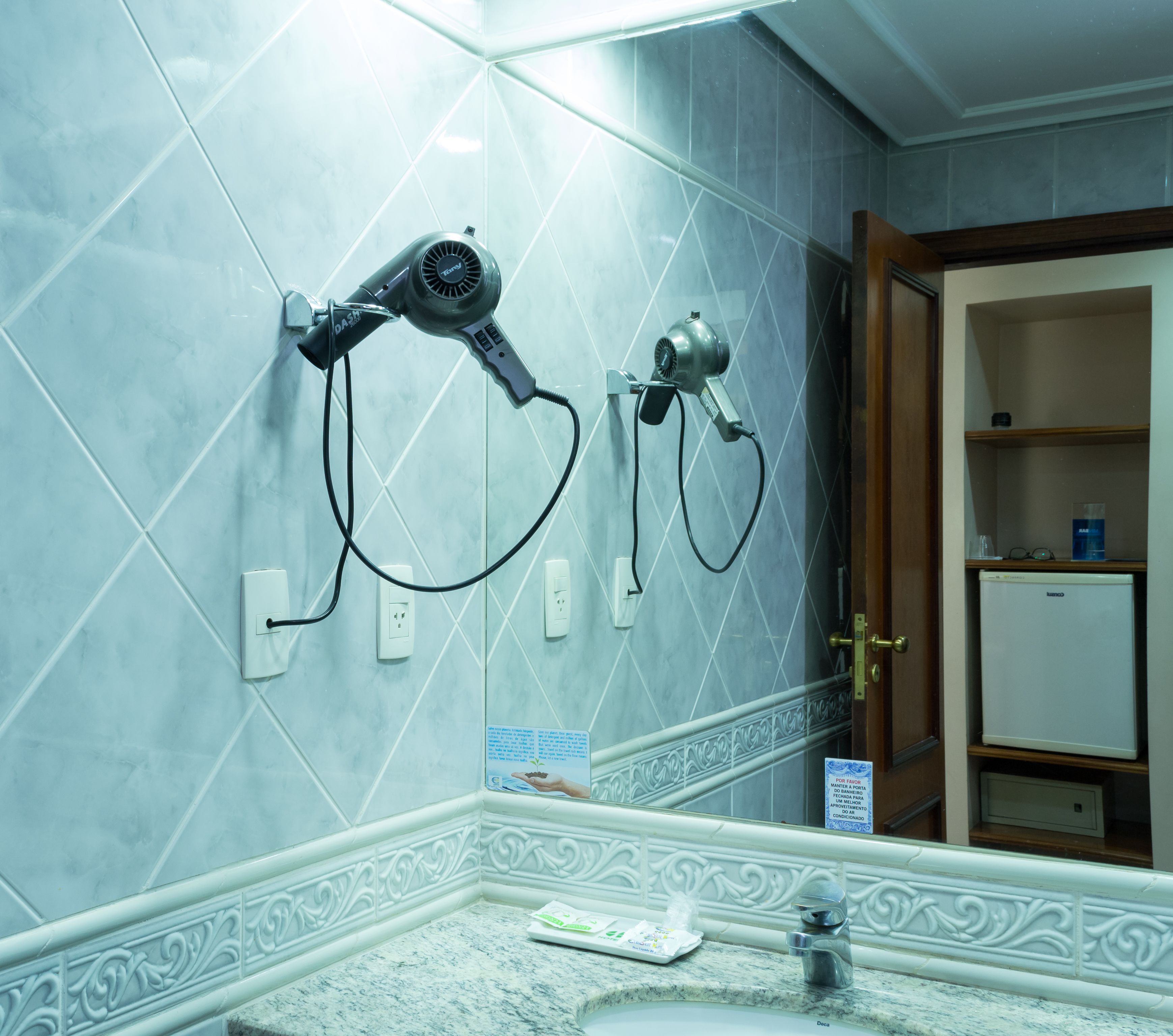 A bathroom with two hair dryers mounted on the wall, a sink with a marble countertop, and a mirror reflecting the bathroom.