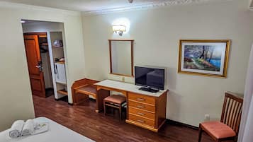 A hotel room with a bed, a desk with a computer, a chair, a small stool, a mirror, and a painting on the wall.