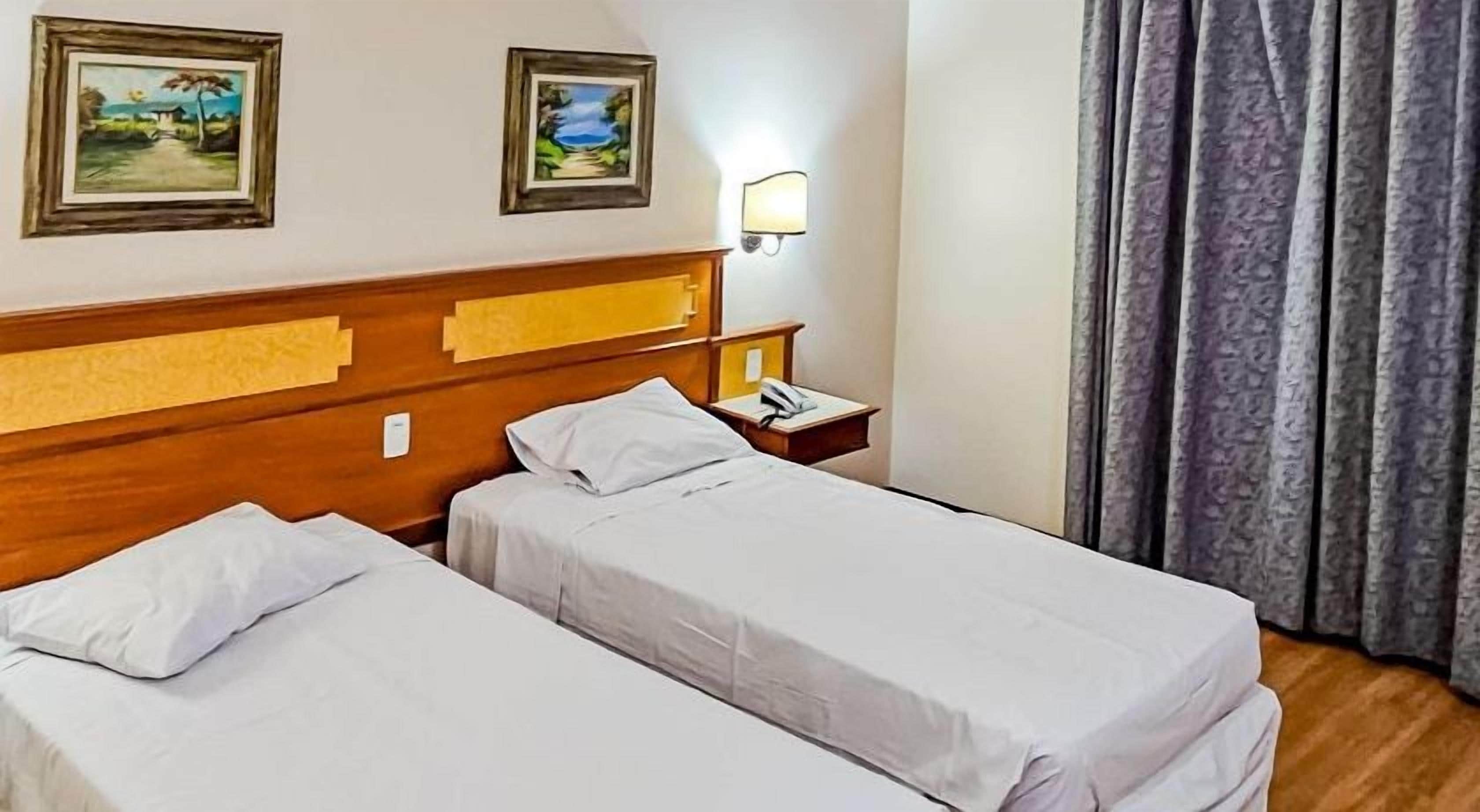A hotel room with two single beds, a wooden headboard, a nightstand with a lamp, and two framed pictures on the wall.