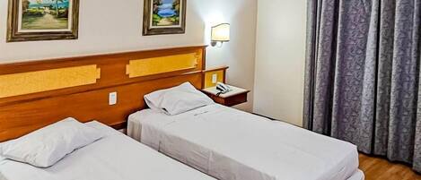 A hotel room with two single beds, a wooden headboard, a nightstand with a lamp, and two framed pictures on the wall.
