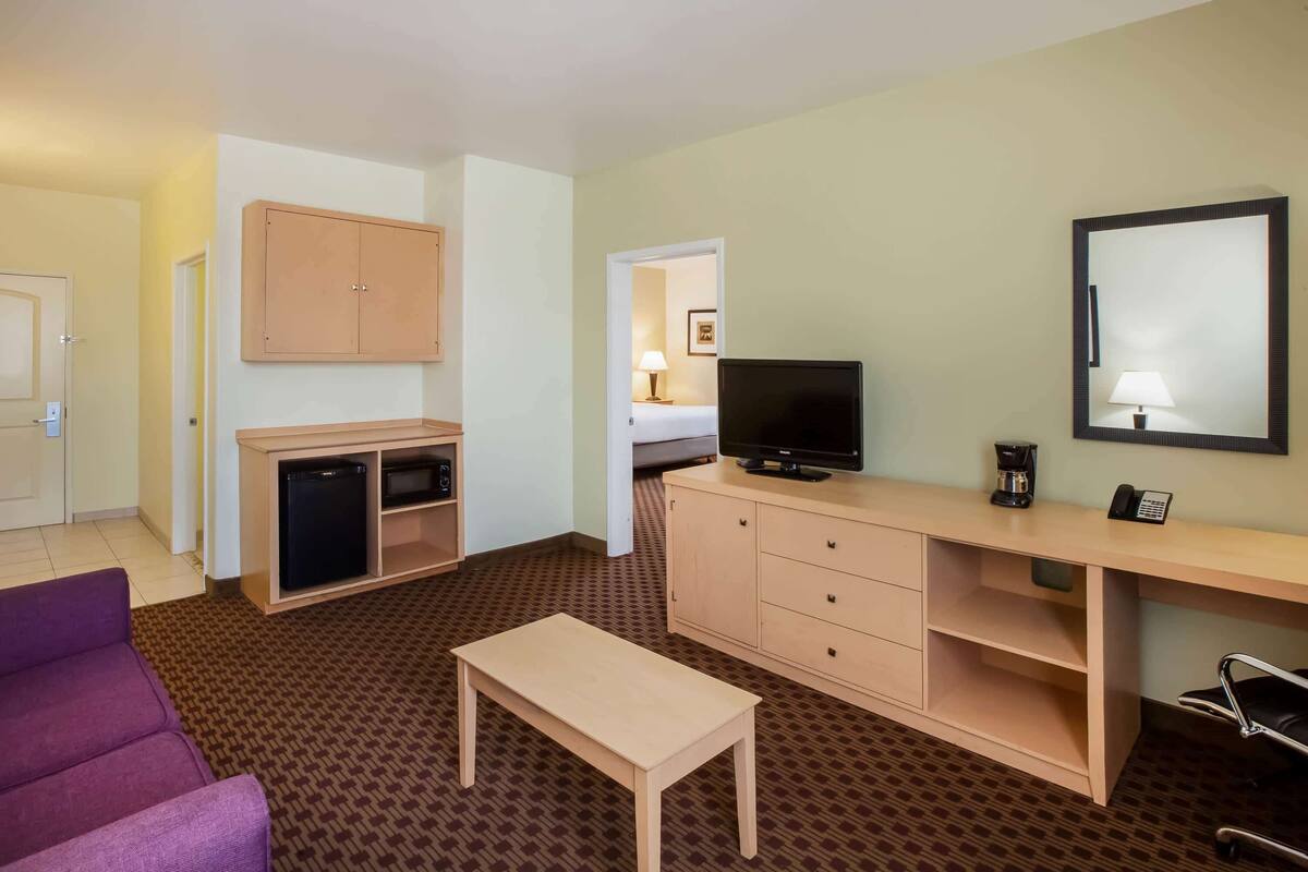Hotelkamer met een bed, een bureau met computer, een televisie, een kleine koelkast, een paarse bank en een houten bankje.