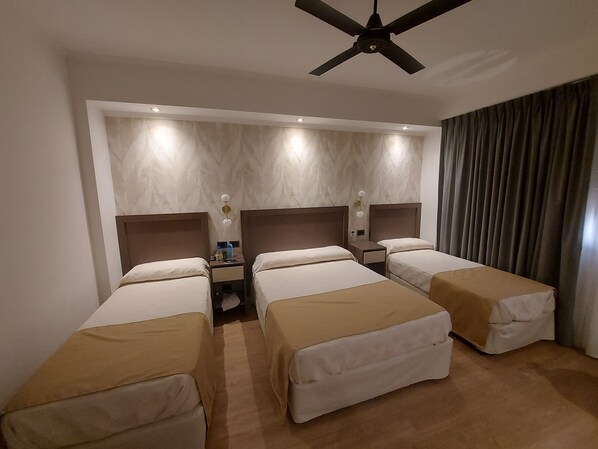 Habitación de hotel con tres camas individuales, un ventilador de techo, una mesita de noche con lámpara y un amplio ventanal con cortinas.