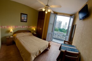 Habitación de hotel con cama, lámparas de noche, escritorio con silla, televisor y balcón con vistas a edificios.