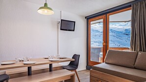 Una habitación acogedora con una mesa de madera dispuesta para cuatro, un sofá cómodo, un televisor montado en la pared y una ventana con vistas a una montaña nevada.