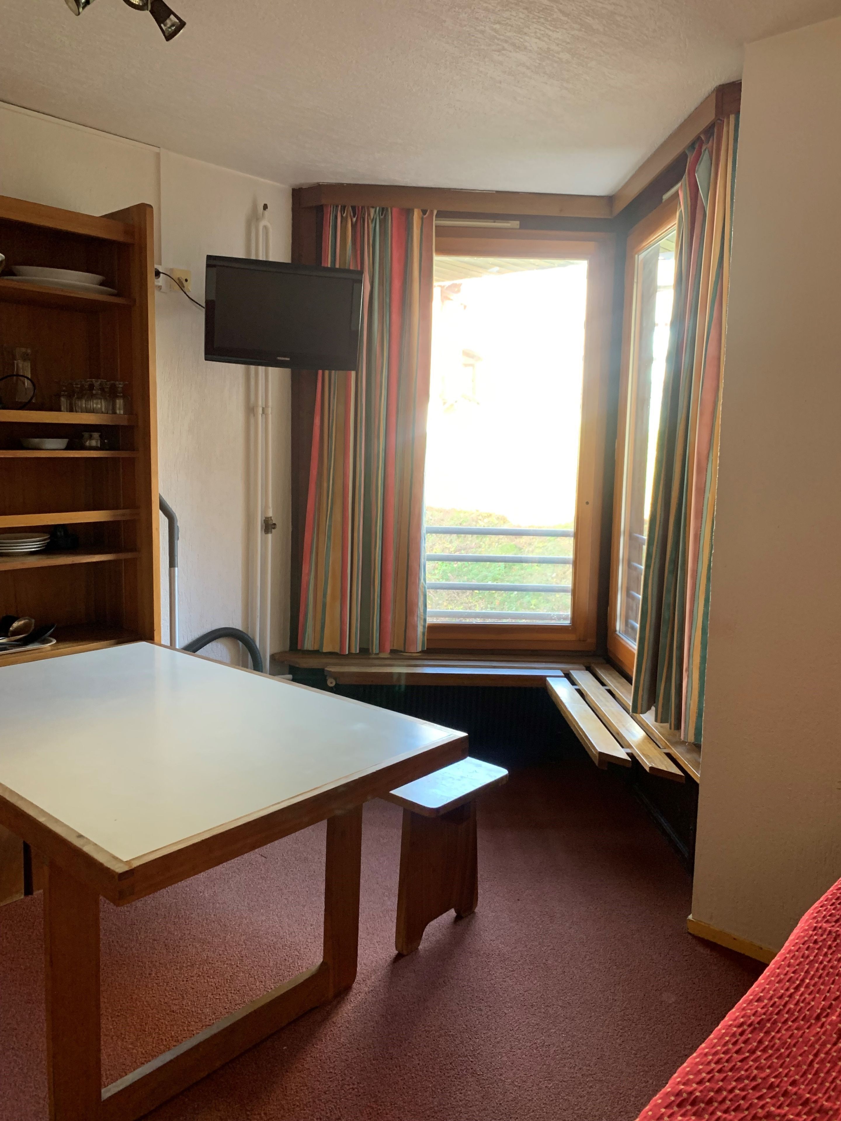Una habitación con una mesa de madera, un banco, un televisor montado en la pared y una ventana con cortinas a rayas.