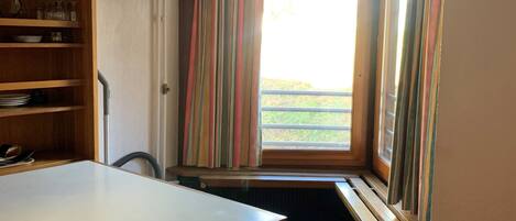 Una habitación con una mesa de madera, un banco, un televisor montado en la pared y una ventana con cortinas a rayas.