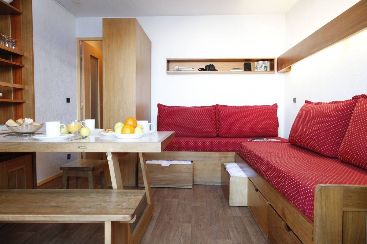 Un espace de vie compact comprenant une table à manger en bois, un banc, un canapé rouge doté de tiroirs de rangement et une petite étagère en bois placée au-dessus du canapé.