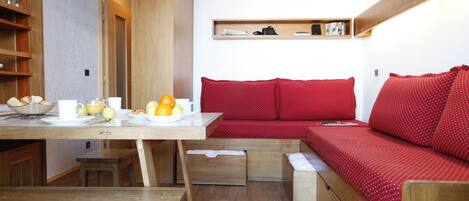 A compact living space with a wooden dining table, a bench, a red sofa with storage drawers, and a small wooden shelf above the sofa.