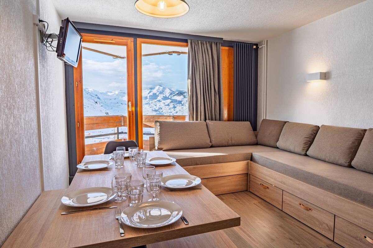 Une pièce moderne comprenant une table à manger en bois pour quatre personnes, un canapé et une grande fenêtre donnant sur les montagnes.
