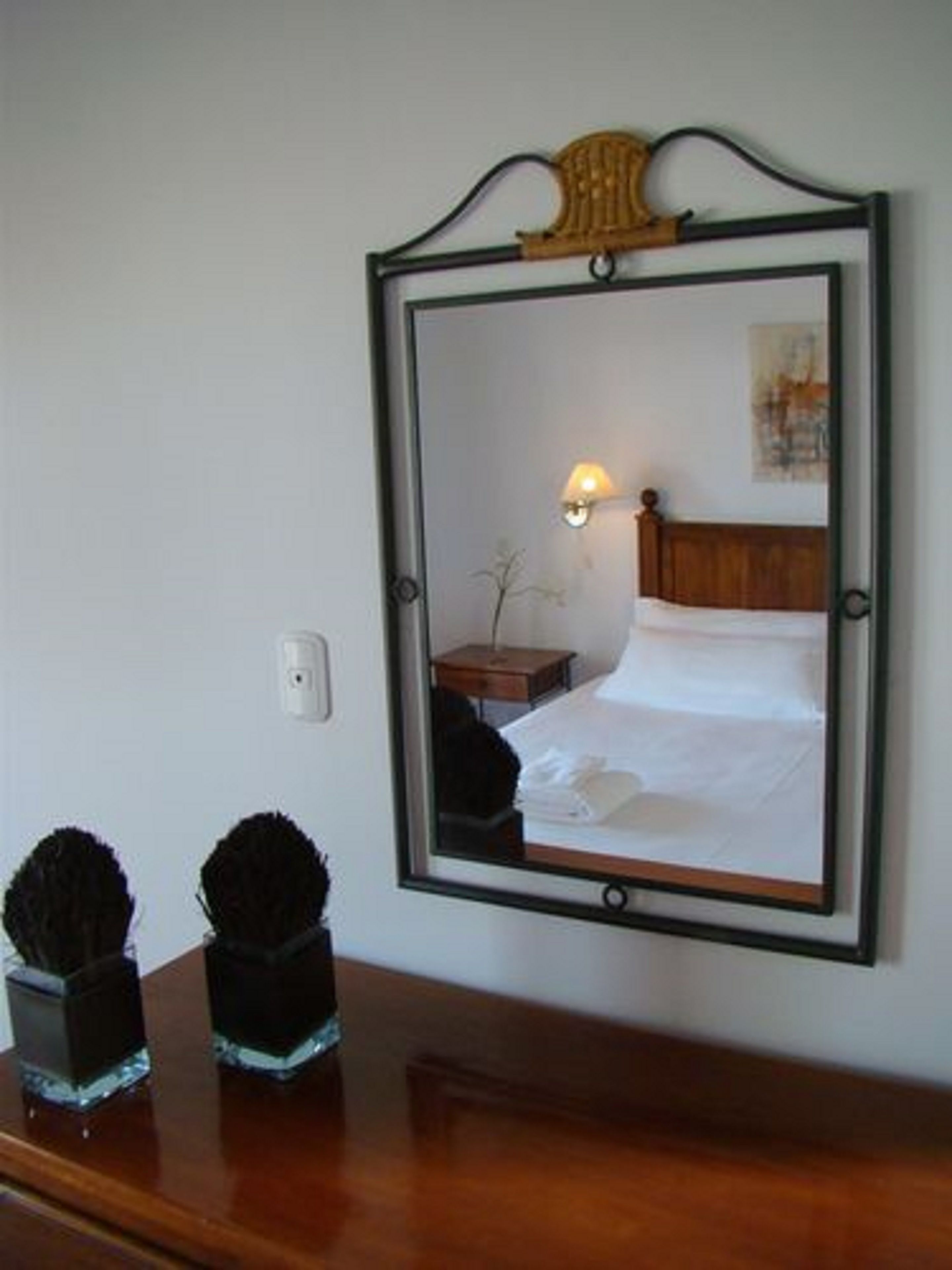 A hotel room with a large mirror, a wooden headboard, a bedside lamp, a small wooden nightstand, and two decorative vases on a wooden dresser.