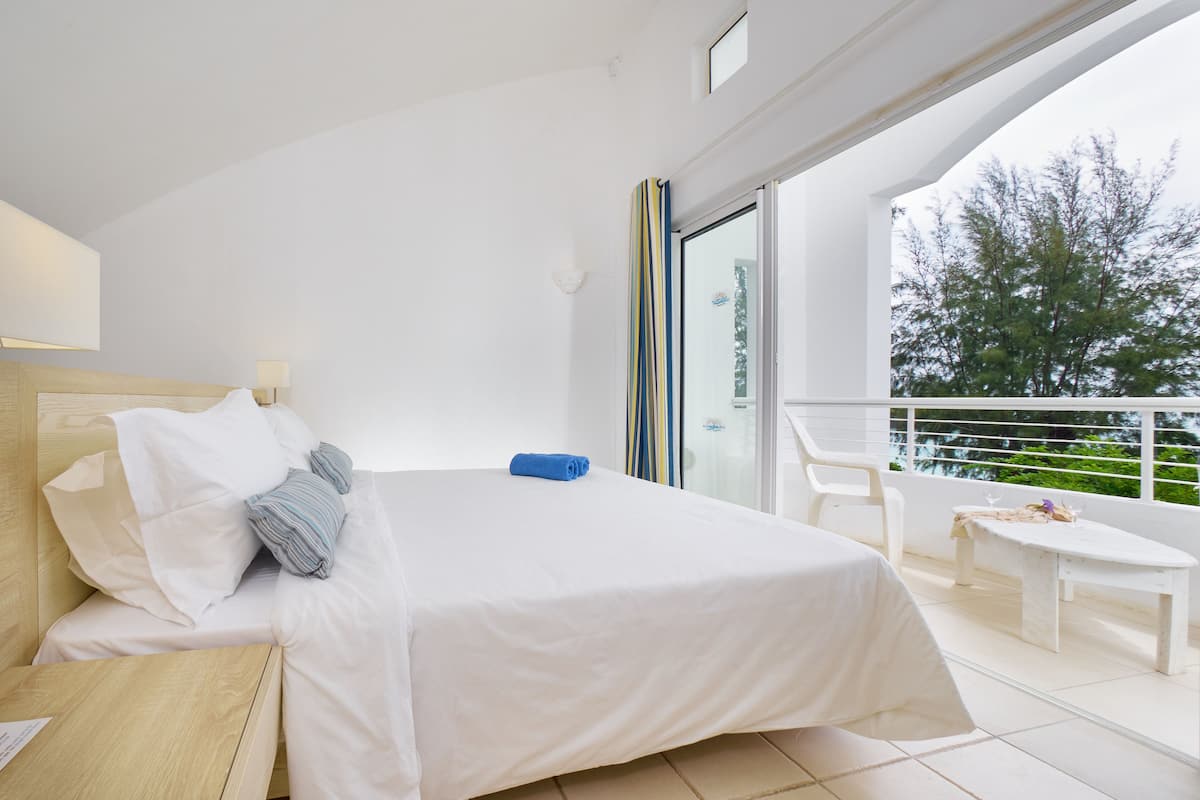 A modern hotel room with a large bed, a wooden nightstand, a small table, and a balcony with a railing.