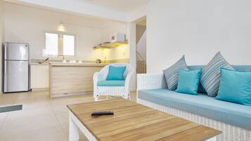 A modern living room with a wooden coffee table, a white sofa with blue cushions, a wicker armchair, and a kitchen area with a refrigerator and microwave.