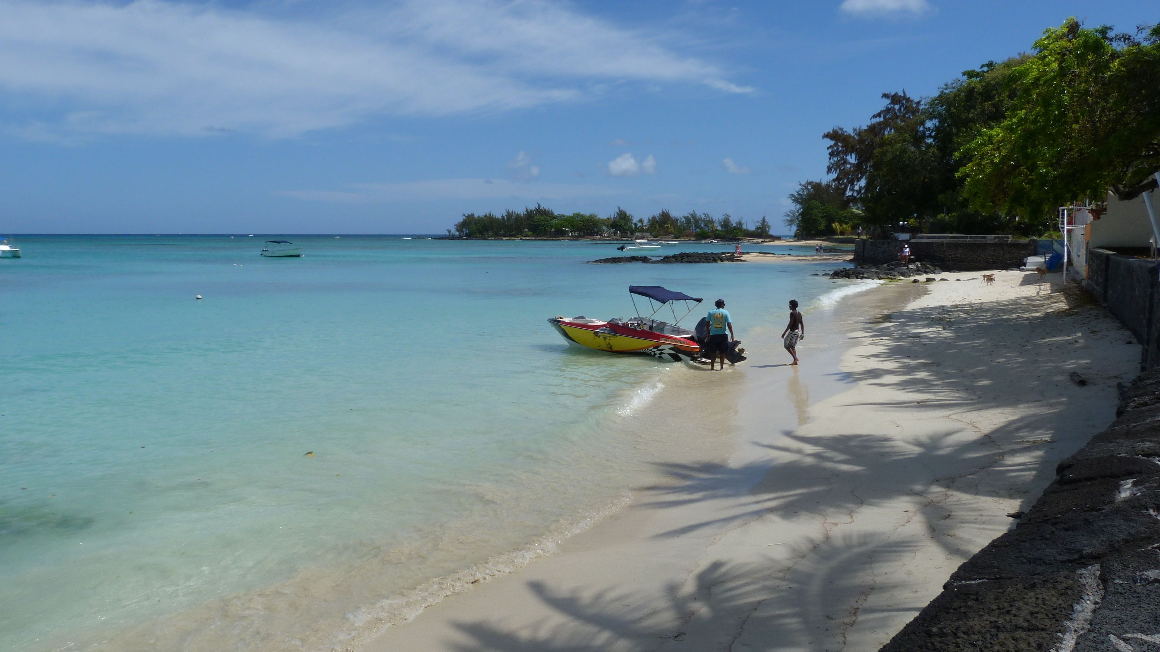 Une plage à l’eau turquoise et limpide, bordée d’une rangée de palmiers, et quelques bateaux.