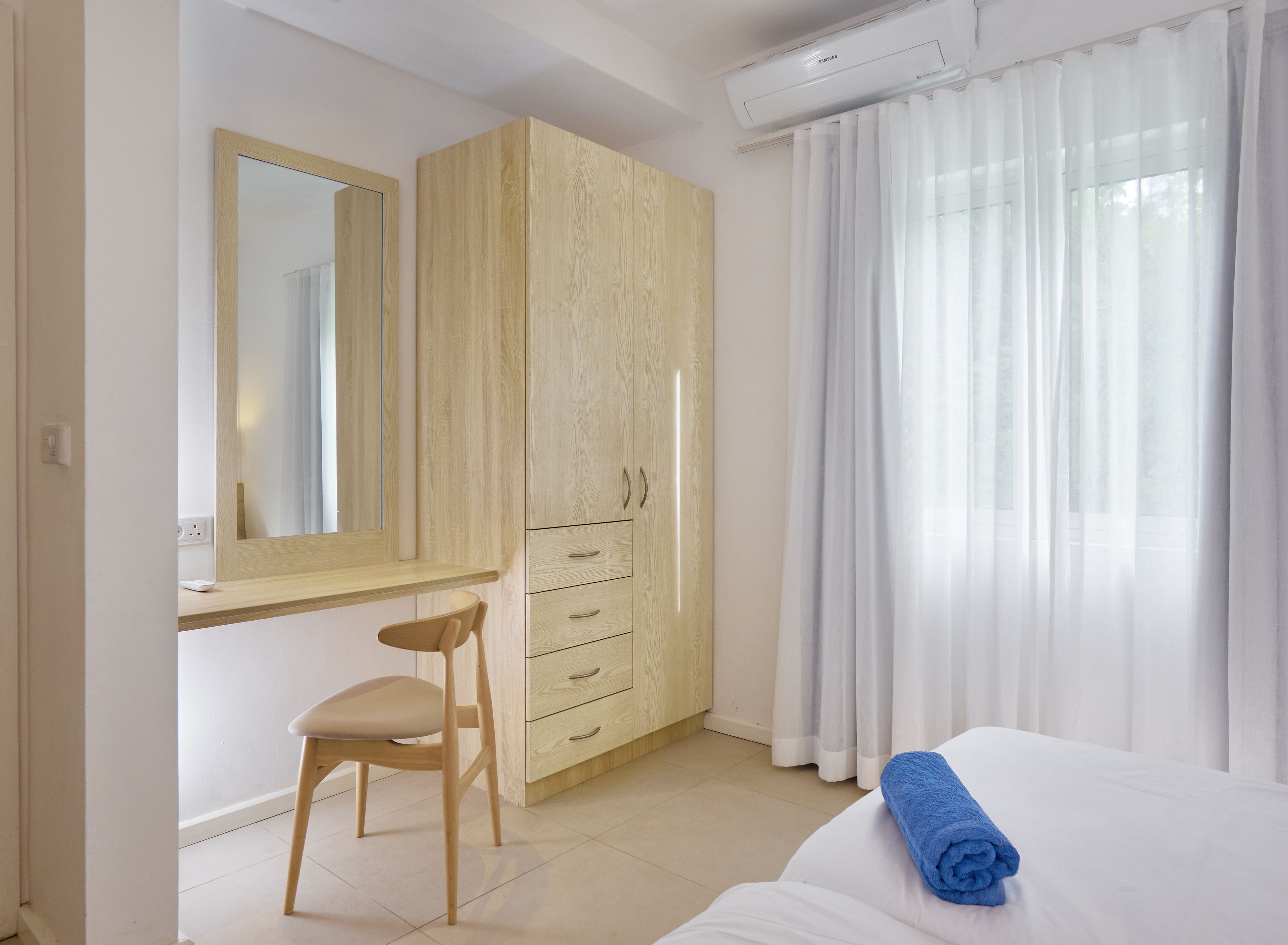 A bedroom with a bed, a wooden wardrobe, a desk with a chair, and a large window with sheer curtains.