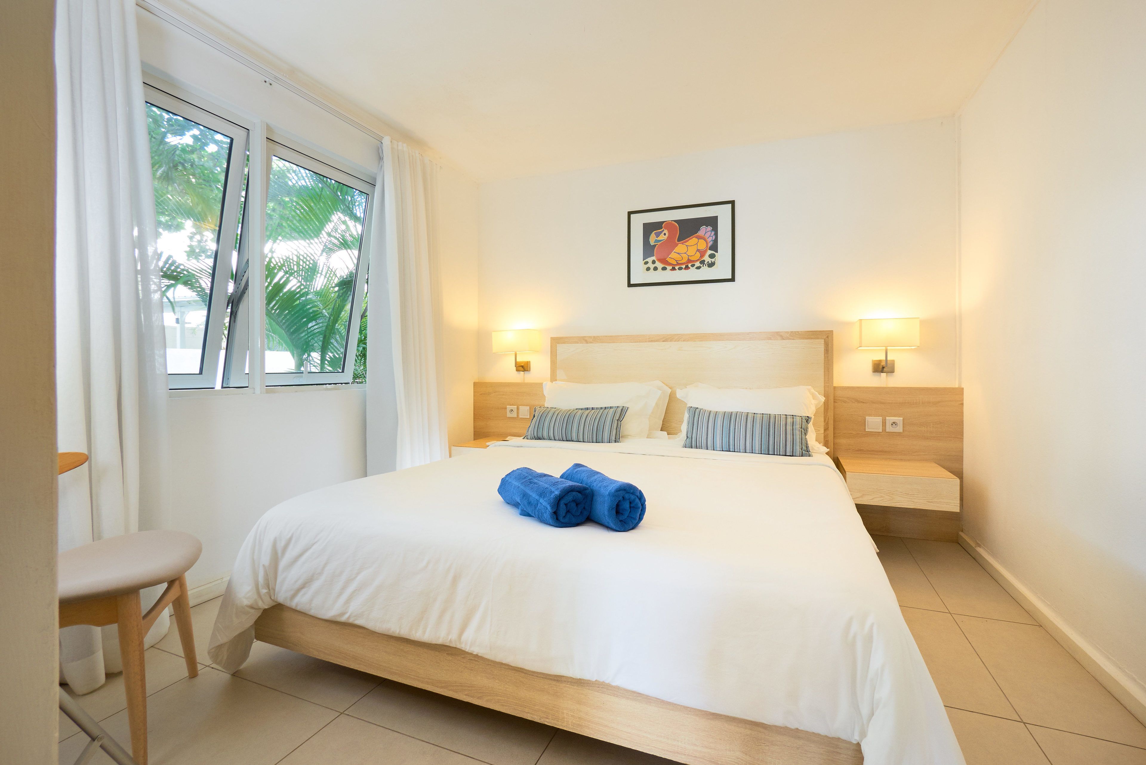 Une chambre d’hôtel avec un grand lit, deux tables de chevet avec des lampes, une fenêtre avec des rideaux et un tableau encadré au mur.