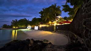 A beach with a stone wall, palm trees, and a building with red umbrellas. - Le Beachclub Serviced Apartments and Villas (Grand-Baie)