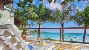 A beachside lounge with white lounge chairs and a small table, overlooking a clear blue sea with boats and palm trees. - Le Beachclub Serviced Apartments and Villas (Grand-Baie)