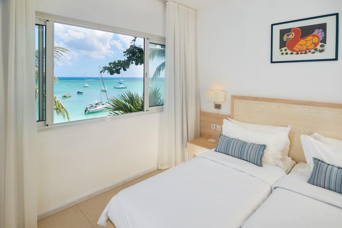 A hotel room with a bed, a nightstand, a lamp, and a window with a view of the sea and boats.