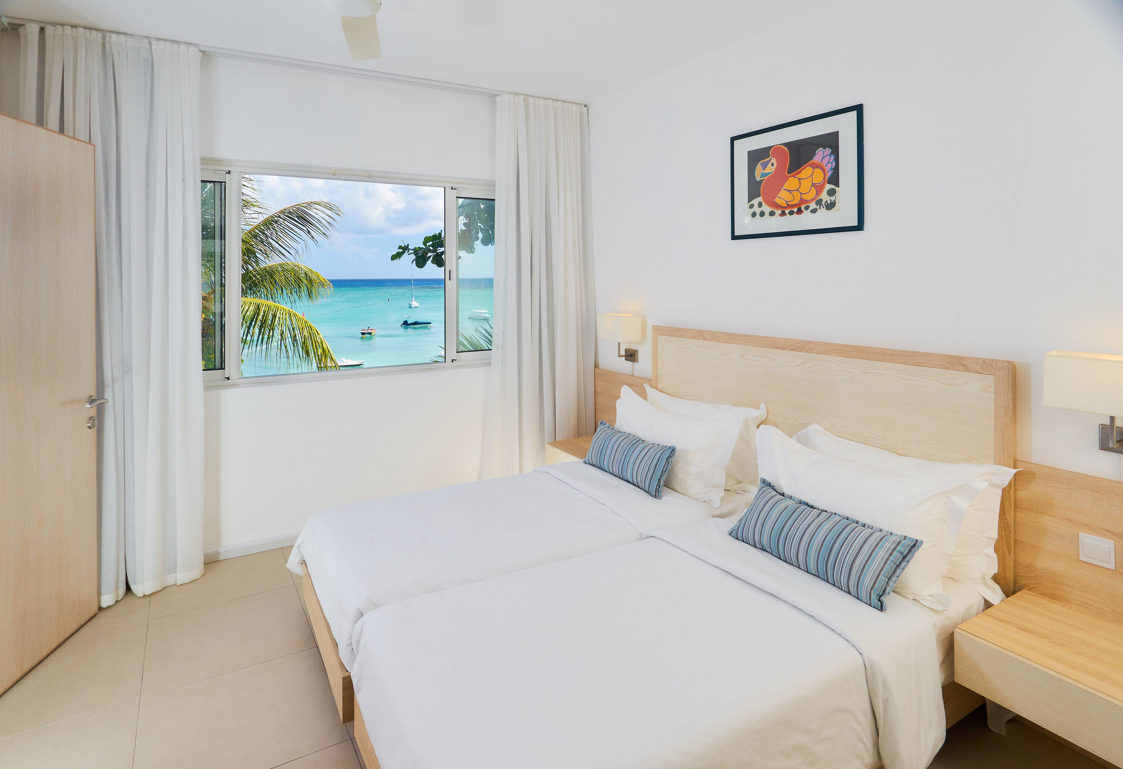 Een hotelkamer met een groot bed, een raam met uitzicht op de oceaan, een houten hoofdbord en een ingelijst beeld aan de muur.