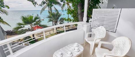 A balcony with white plastic chairs and a table, overlooking a beach with palm trees and boats.