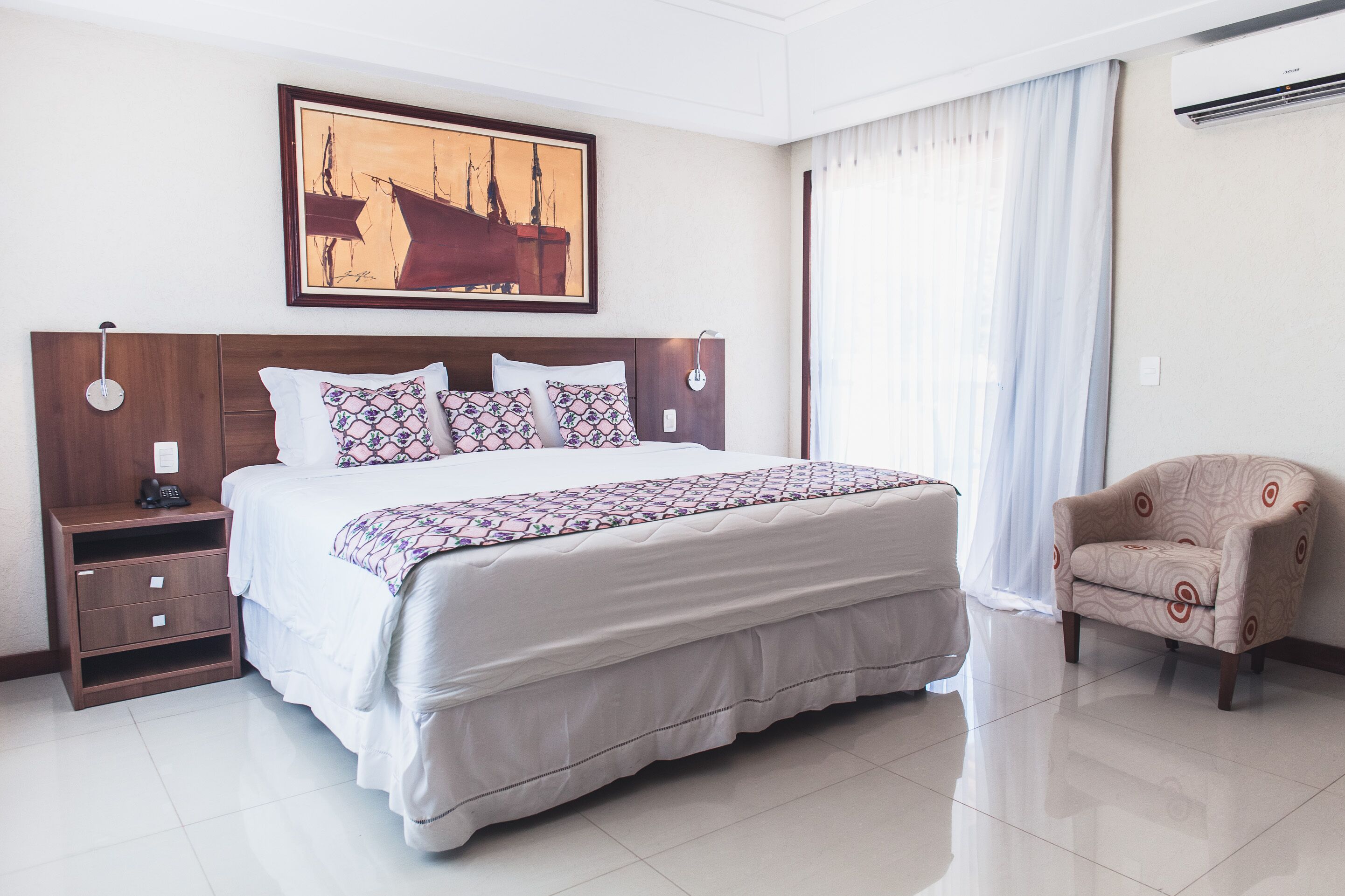 A hotel room with a large bed, a nightstand, a chair, a painting on the wall, and a window with curtains.