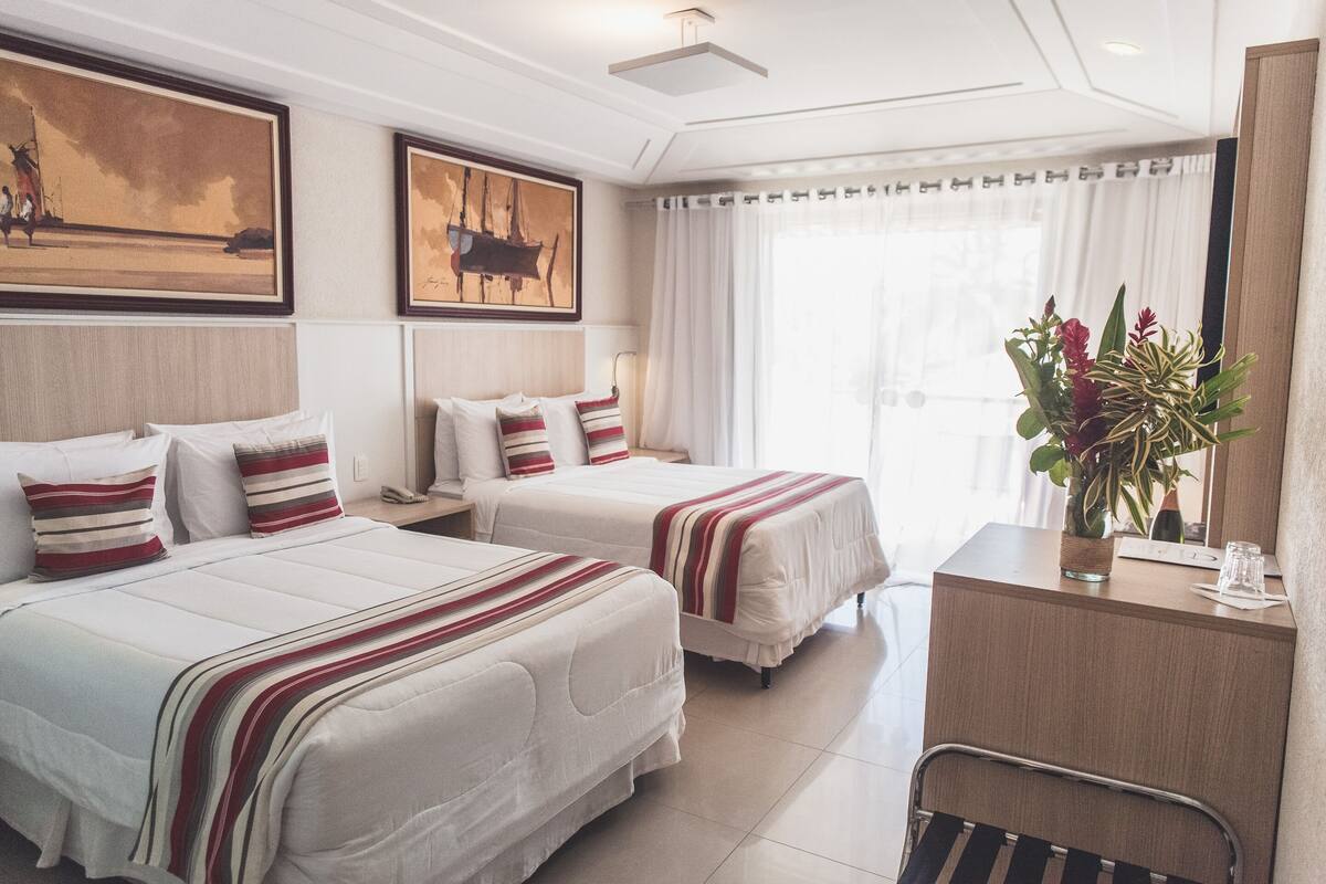 Une chambre d’hôtel avec deux lits, un bureau, une chaise et une grande fenêtre avec des rideaux.