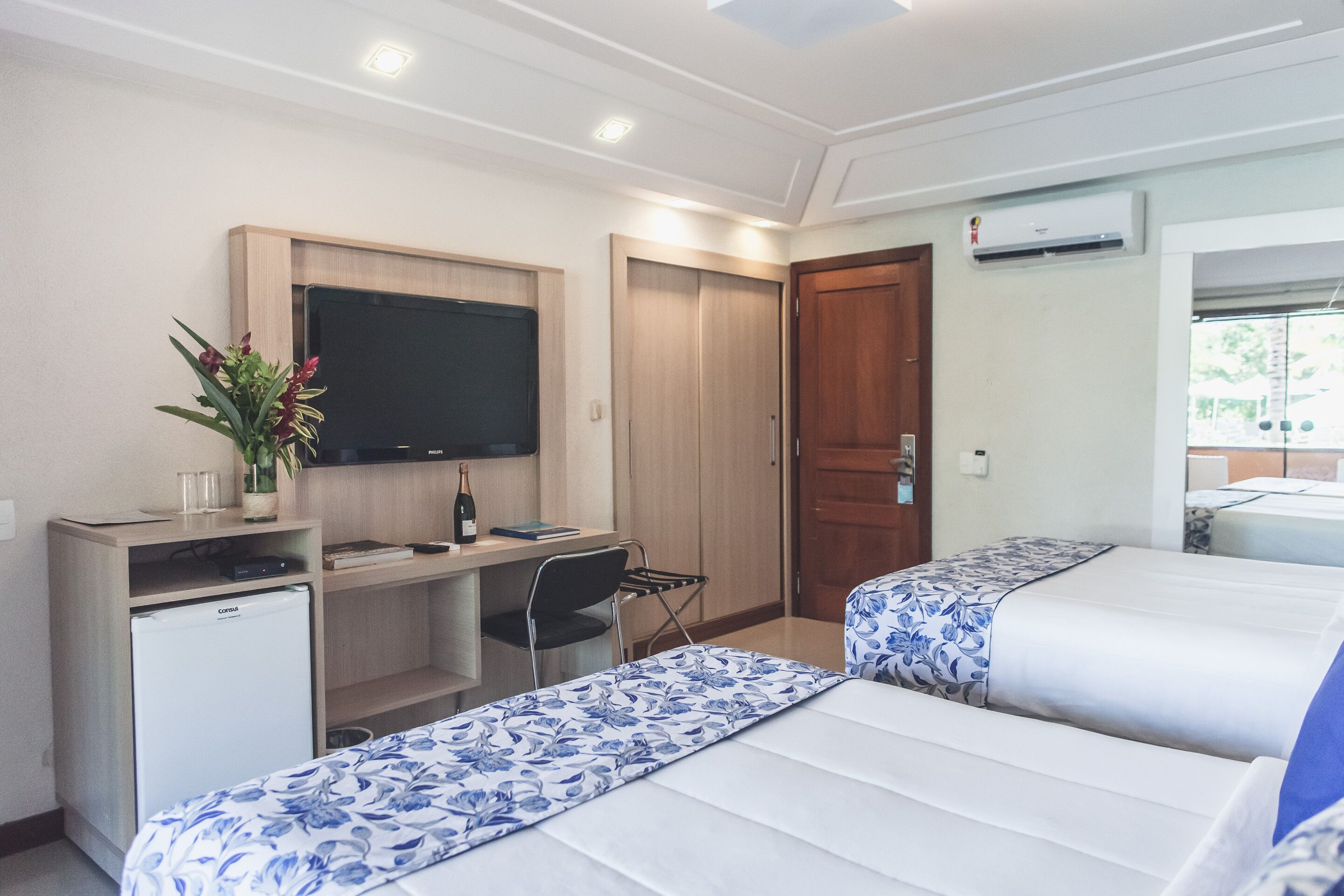 A hotel room with two beds, a desk, a television, a small refrigerator, and an air conditioning unit.