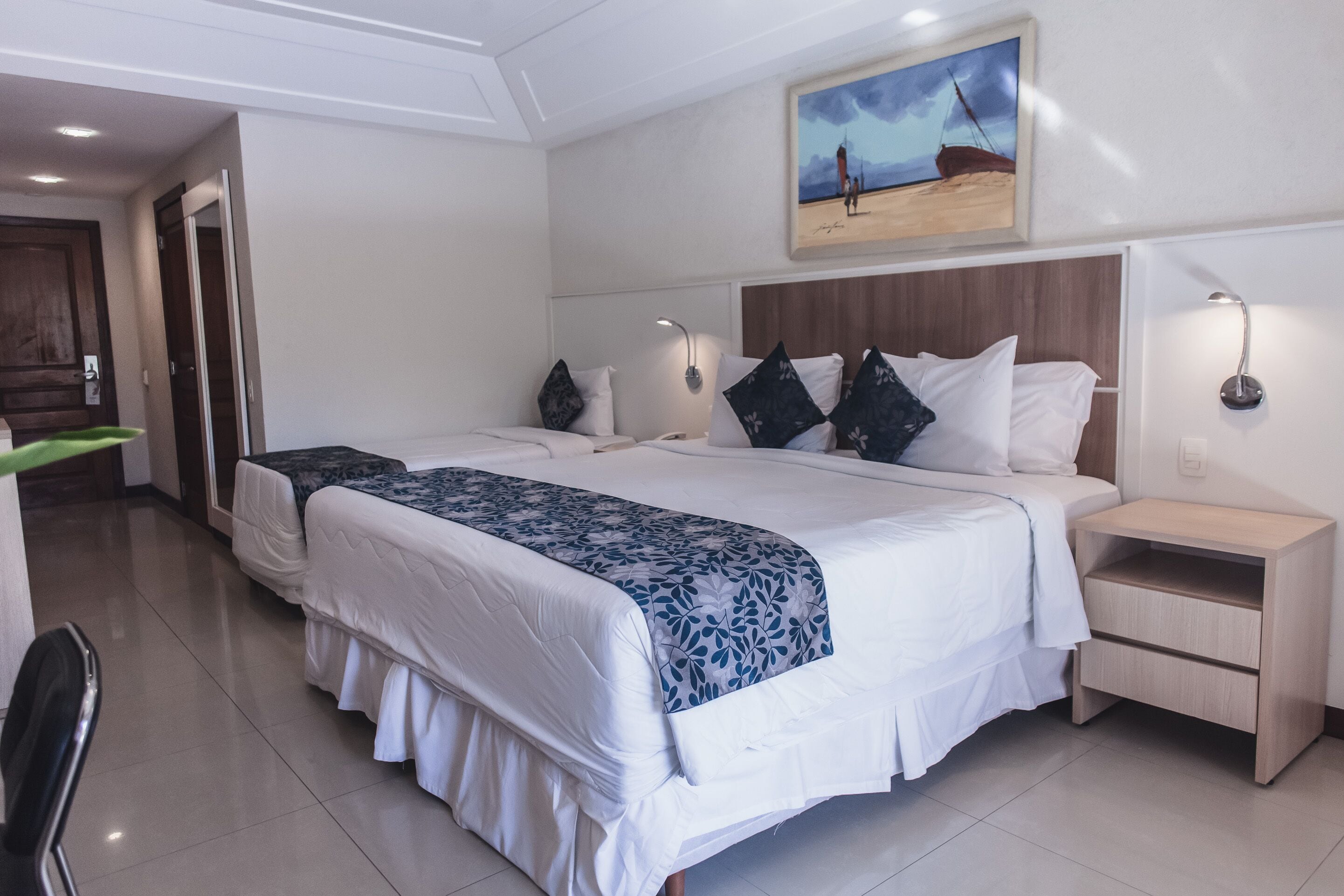 A hotel room with two beds, a wooden headboard, bedside lamps, a painting on the wall, and a door leading to another room.