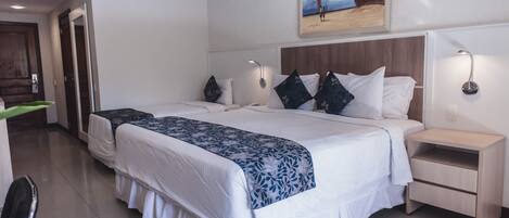 A hotel room with two beds, a wooden headboard, bedside lamps, a painting on the wall, and a door leading to another room.