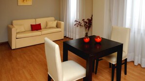 A living room with a beige sofa, a dark wooden table with two chairs, and a vase with red flowers. - Del Bono Suites Art Hotel (San Juan)