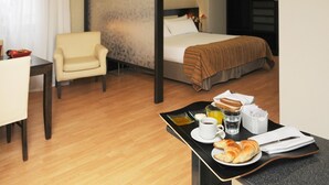 A hotel room with a bed, a chair, a dining table with breakfast items, and a view of the city through the window. - Del Bono Suites Art Hotel (San Juan)