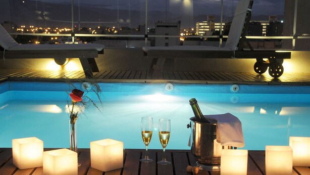 A rooftop pool area with a wooden deck, two glasses of wine, a bottle of champagne, a vase with a single flower, and several lit candles. - Del Bono Suites Art Hotel (San Juan)