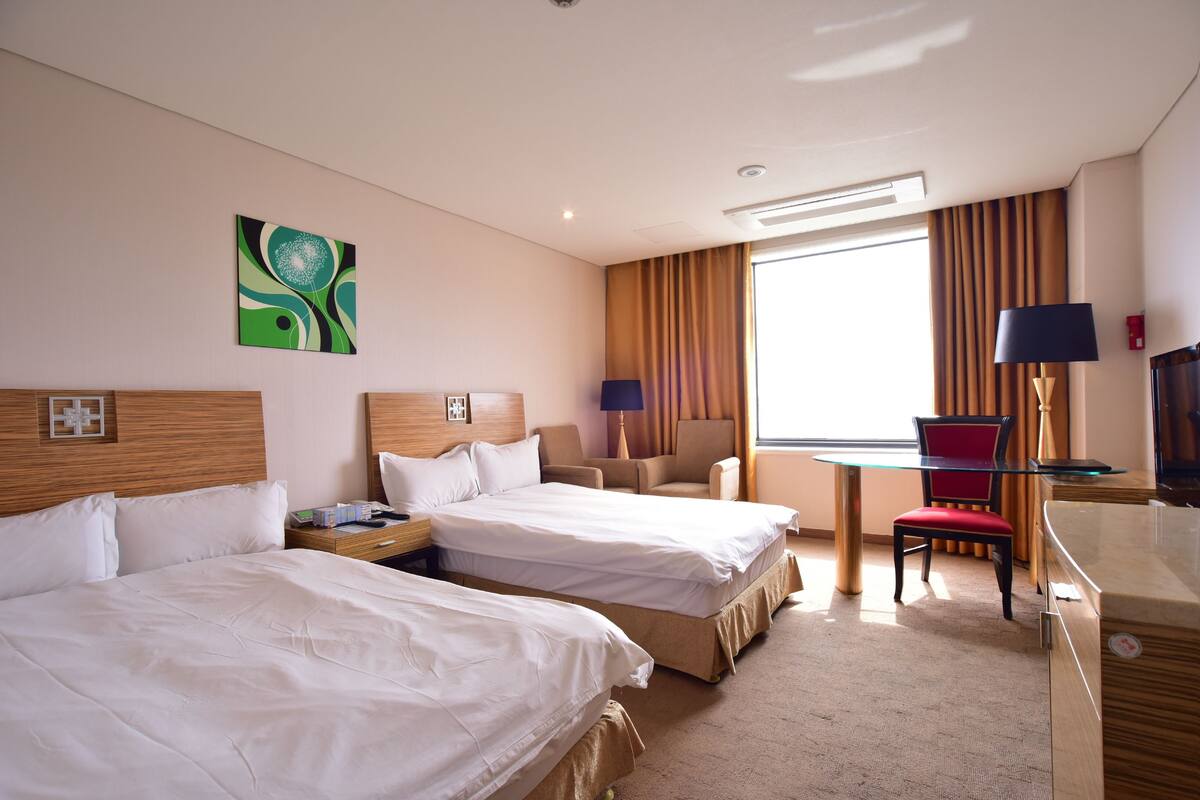 A hotel room with two beds, a desk with a chair, a TV, and a large window with curtains.