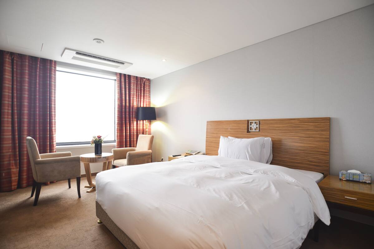 A hotel room with a large bed, a desk, a chair, a lamp, and a window with curtains.