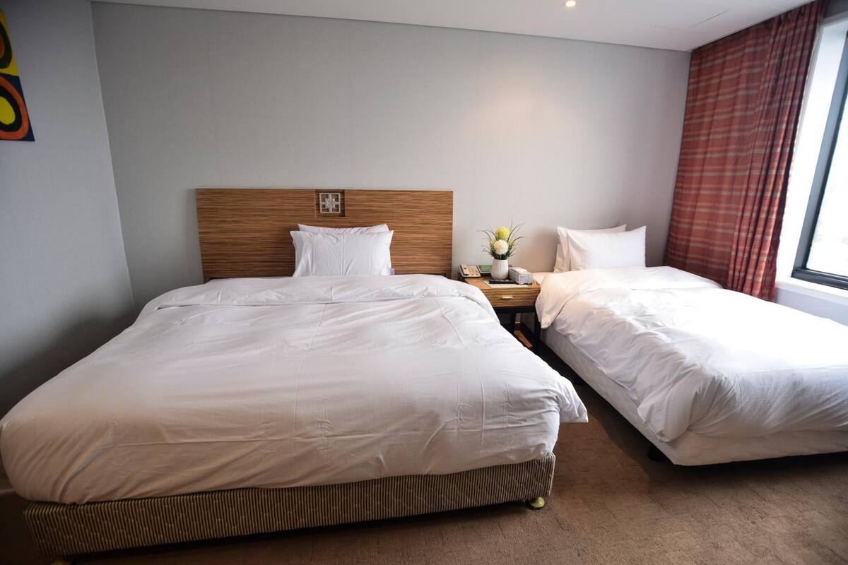 A hotel room with two beds, a wooden headboard, a nightstand with a phone and a vase of flowers, and a window with curtains.