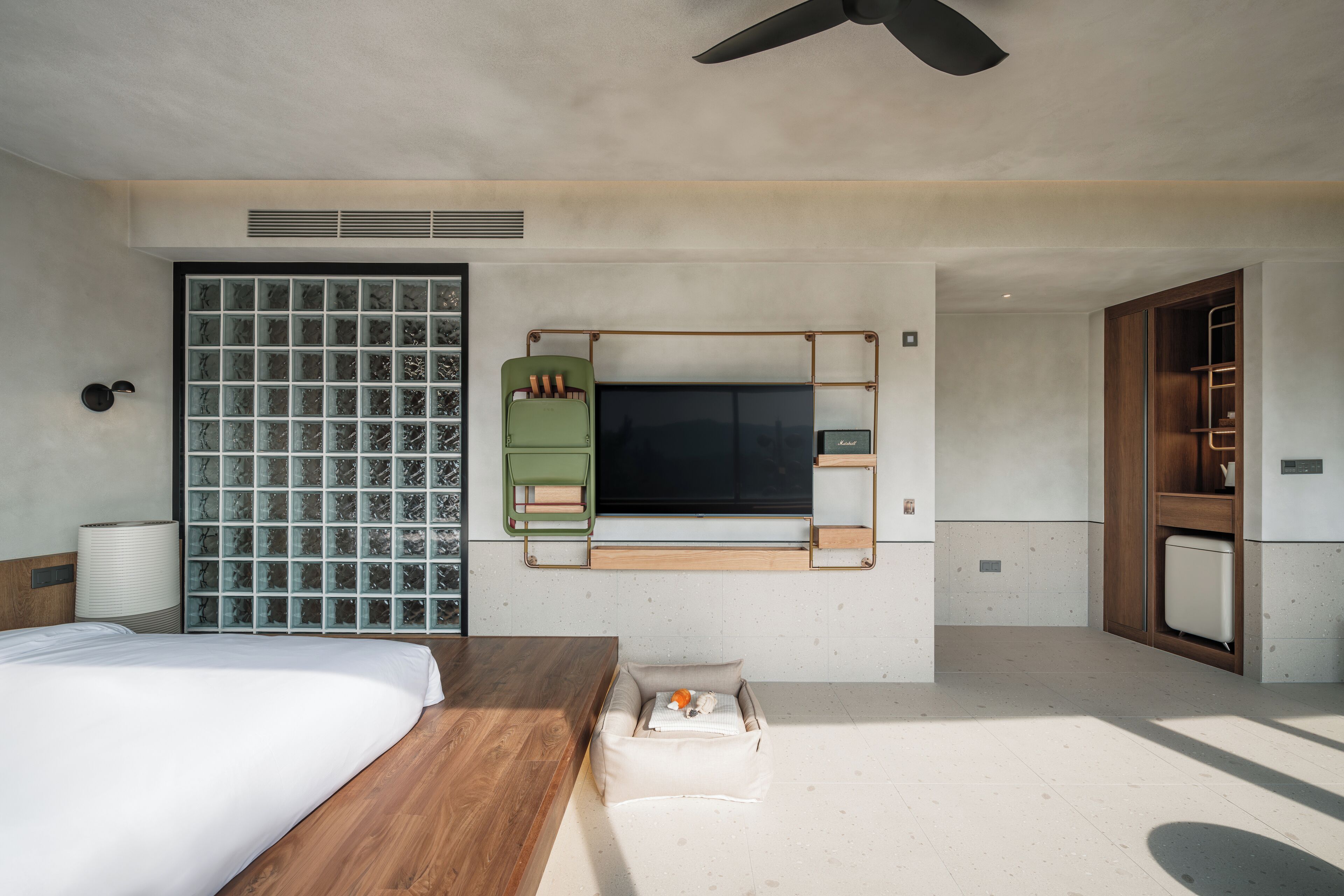 A modern hotel room with a large bed, a wooden side table, a built-in TV, a green chair, and a large window with a view of the city.