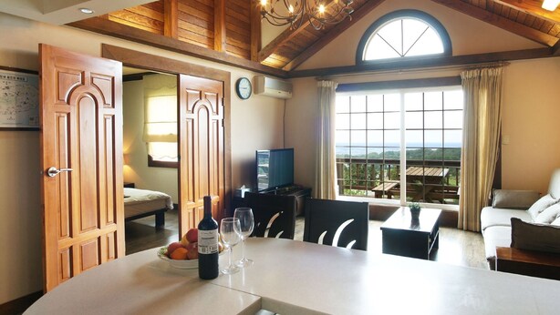 A dining area with a table, chairs, a bottle of wine, and a bowl of fruit. There is a sofa and a television in the background. - Aewol ParkHill Village (Jeju City)